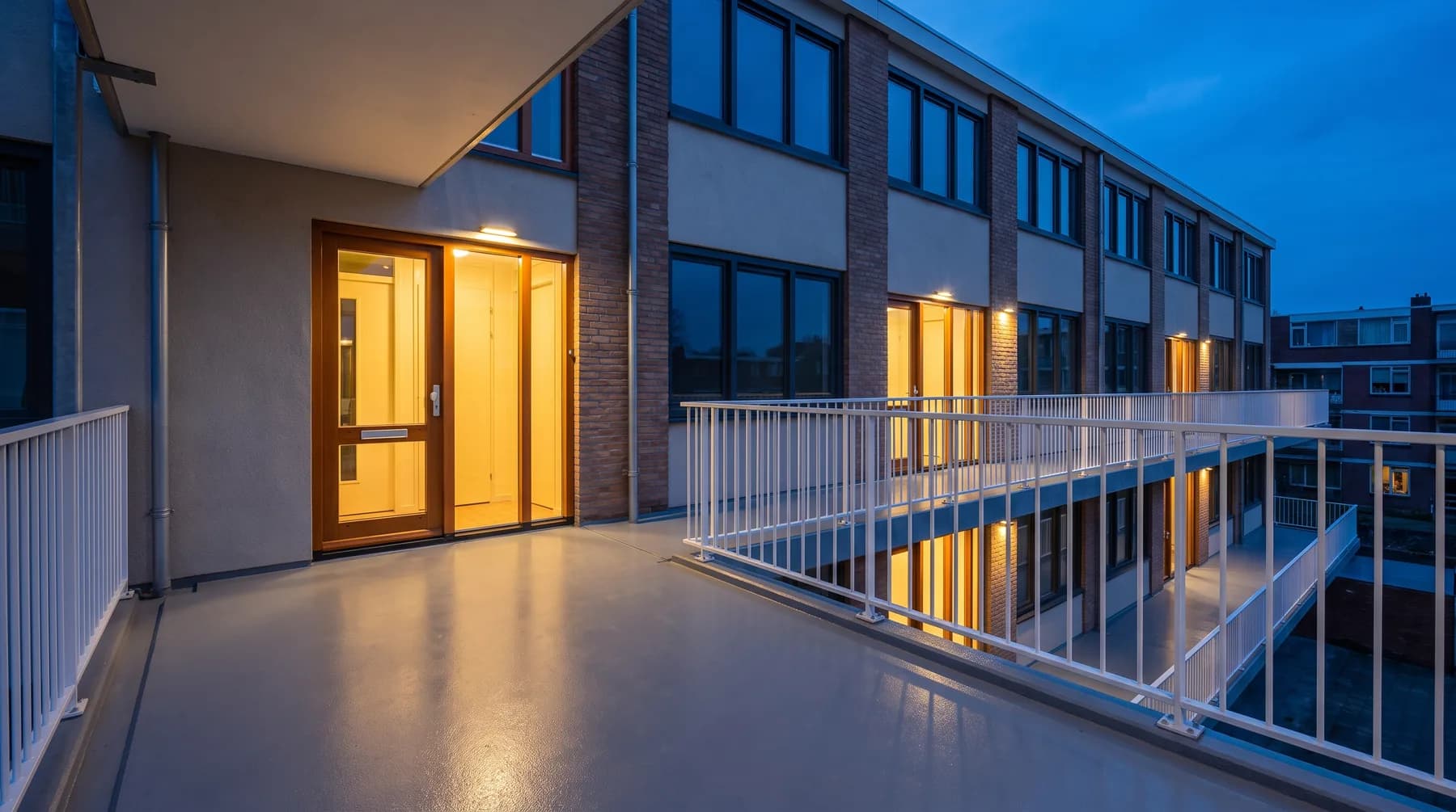 Galerij van een appartementengebouw in het blauwe uur, met warm licht uit de voordeuren